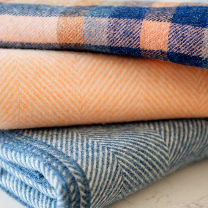 Stack of folded blankets, Falmouth Blue and Ivory Check, Blueberry Swirl and Mango Sorbet Herringbone Shetland Wool Blankets on a marble surface 