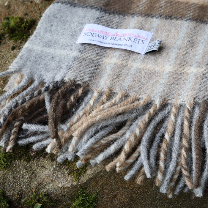 McKellar Tartan Plaid blanket, in soft grey, oatmeal and brown with fringes on a textured surface outdoors, featuring a Solway Blankets label. 