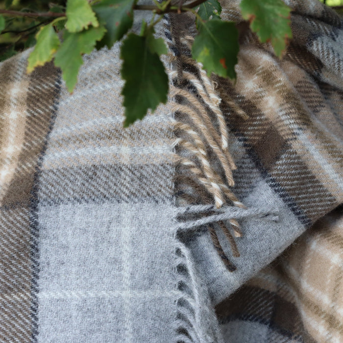 Natural coloured McKellar tartan knee blanket in grey, oatmeal and brown tones, draped over a branch with green leaves in the background