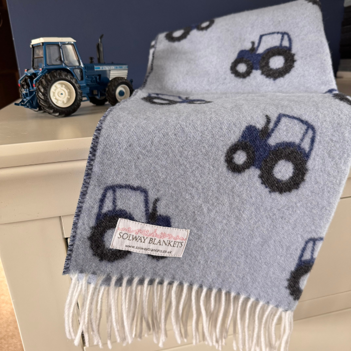 Blue Tractor patterned merino wool baby blanket with fringe and a repeating tractor pattern, draped over a white cabinet next to a blue toy tractor. A brand label reading 'SOLWAY BLANKETS' is visible on the blanket.