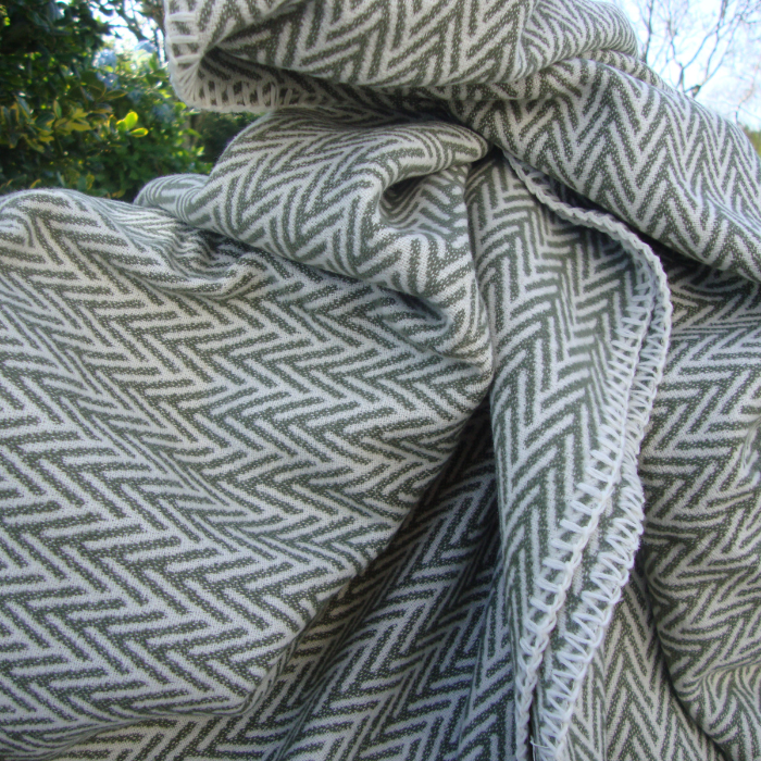 A close-up of a woven throw blanket with a mineral green and off-white herringbone pattern, draped outdoors with natural light and foliage visible in the background.