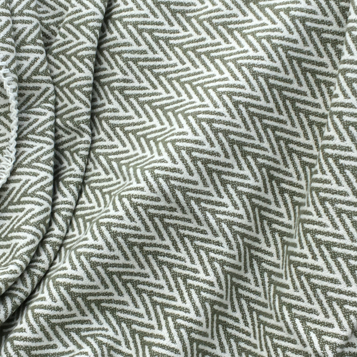 A close-up of a woven Mineral Green cotton throw with a herringbone pattern in olive green and off-white, showing the fabric texture and folds.