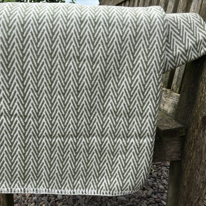 A woven throw blanket with a mineral green and off-white herringbone pattern draped over a rustic wooden bench arm outdoors.