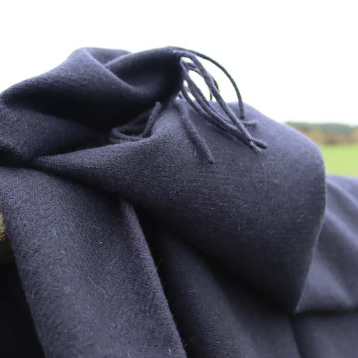 A close-up of a dark navy cashmere wool throw with delicate fringed edges, draped in soft folds against a bright outdoor background.