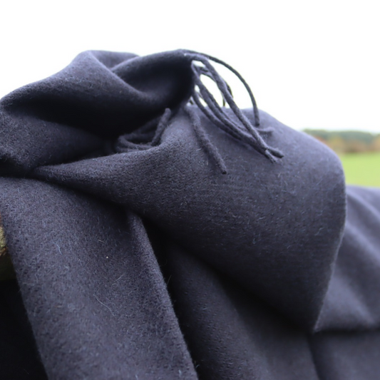 A close-up of a dark navy cashmere wool throw with delicate fringed edges, draped in soft folds against a bright outdoor background.
