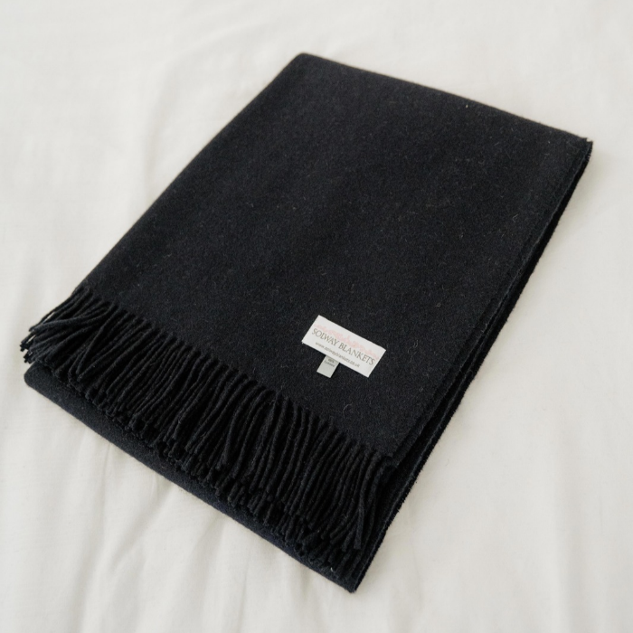 Close-up of the fine weave and soft texture of navy Scottish cashmere with fringe detailing, neatly folded and a small white brand label reading 'Solway Blankets', displayed on a white fabric background.