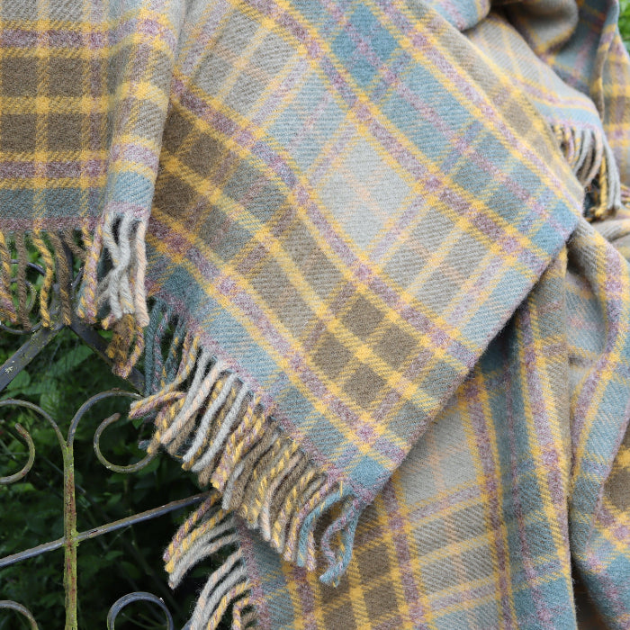 Olive green, teal blue and mustard wool blanket draped over iron gate outdoors with trees and green shrubbery in the background