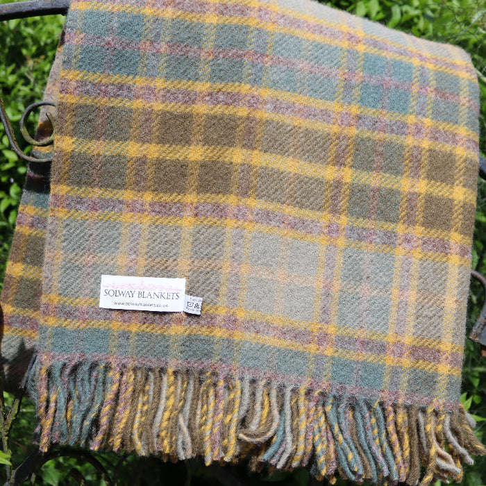 Olive green, teal blue and mustard wool blanket with Solway Blankets label folded neatly over iron gate with trees and green shrubbery in the background