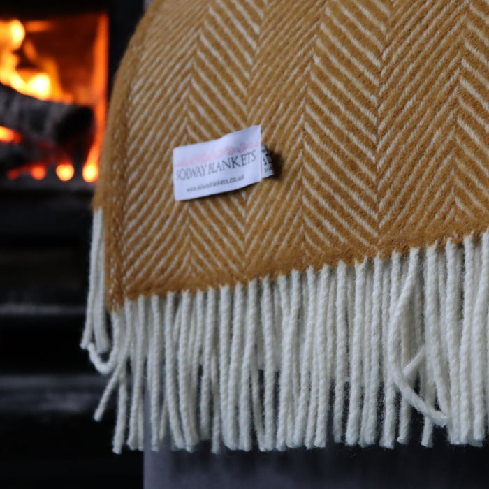 Mustard coloured woven wool blanket with fringe and a Solway Blankets label in front of a cosy fireplace