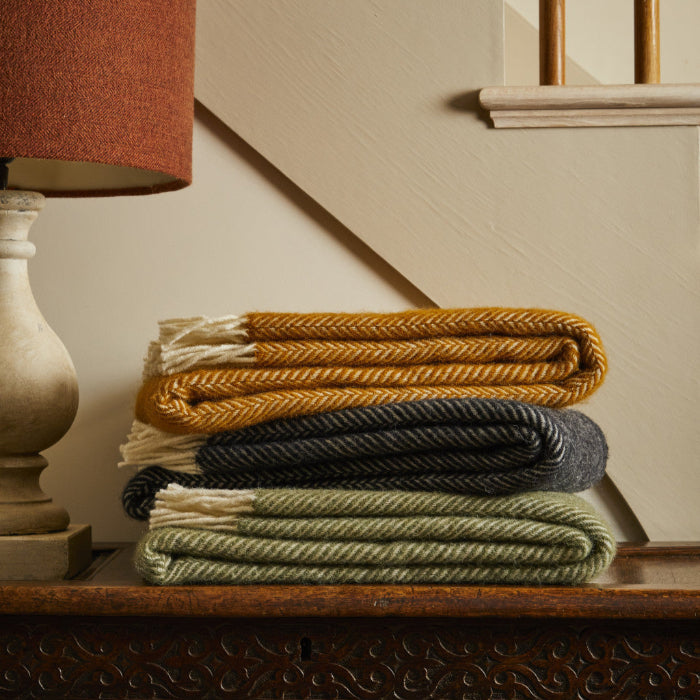 Stack of folded wool blankets in yellow, charcoal, and green on a wooden dresser with an orange lamp in the background.