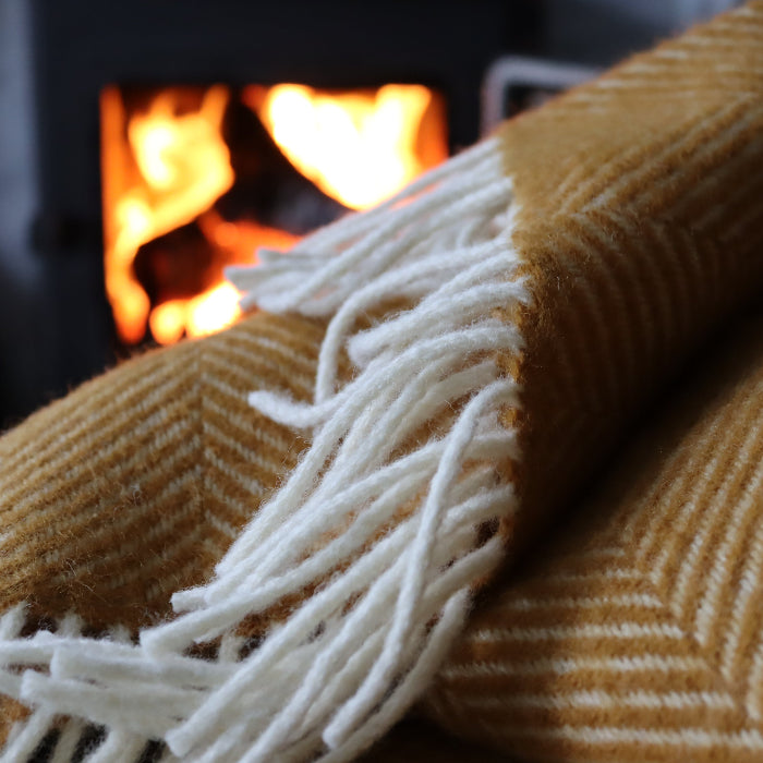Close up of a mustard fishbone wool blanket with white fringe, with a cosy fireplace in the background