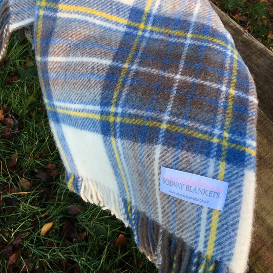 Muted Blue Tartan wool blanket throw with fringe, featuring a Solway Blankets label, draped over a wooden bench outdoors on a grassy ground