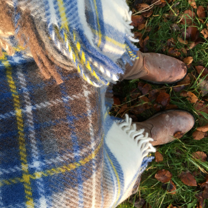 Person with Muted Blue tartan wool knee blanket wrapped around them, wearing brown boots on grass with fallen leaves