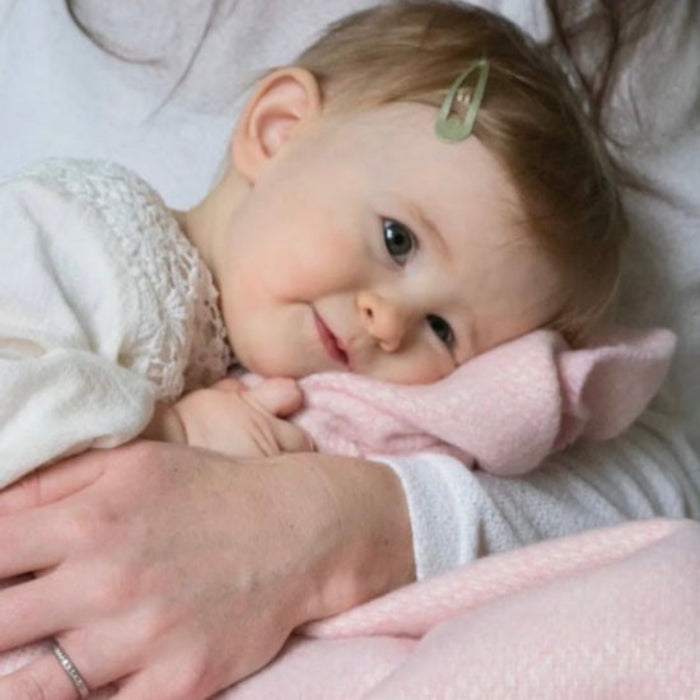 Baby being cuddled and holding the Niamh Pink Cashmere Baby Blanket with a blurred background