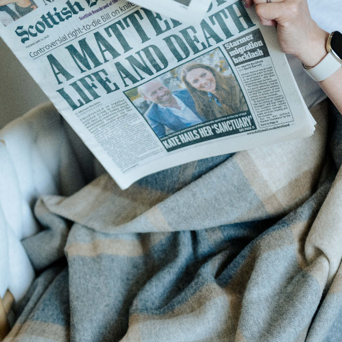 Person reading a newspaper titled 'A Matter of Life and Death' wrapped up in a natural blanket made up of oatmeal, biscuit and shades of grey