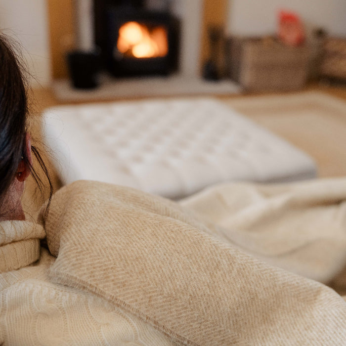 Person wrapped in a Natural Herringbone Shetland Wool Throw in a cosy room with a fireplace.