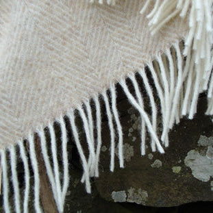 Close-up of a beige Natural Herringbone Shetland Wool woven blanket with white fringe on a textured surface.