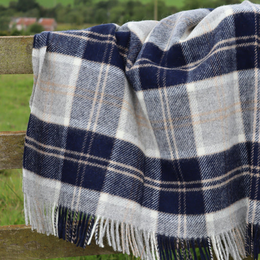 Navy, white and beige plaid blanket throw with fringes draped over a wooden fence in a grassy field.