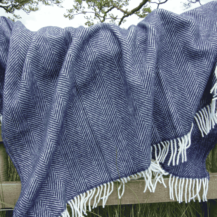 Navy blue fishbone wool blanket with white fringe, draped over a wooden fence in a grassy background