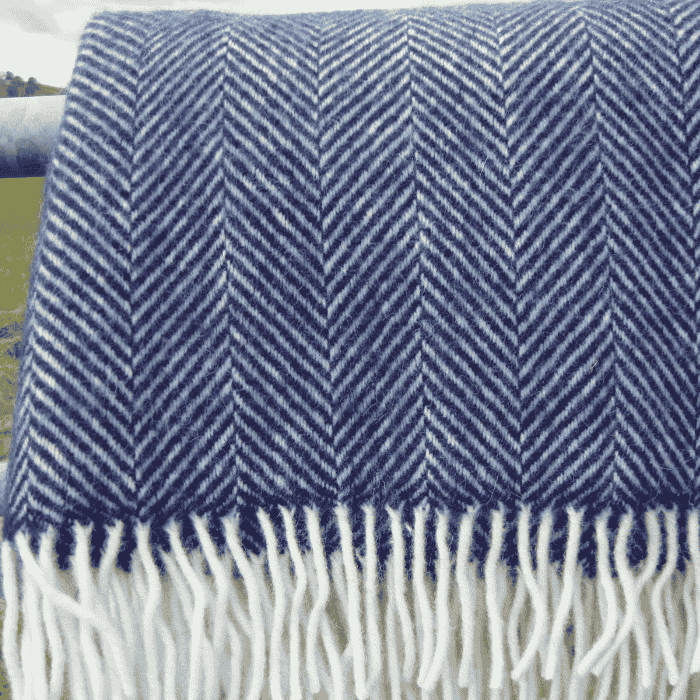 Up close of a navy blue fishbone wool blanket with white fringe, draped over a metal fence in a grassy field.