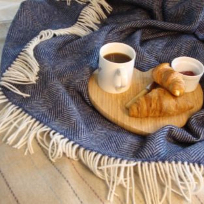 Navy blue herringbone blanket throw with white fringe under a wooden tray holding coffee and pastries, on a light wooden floor.