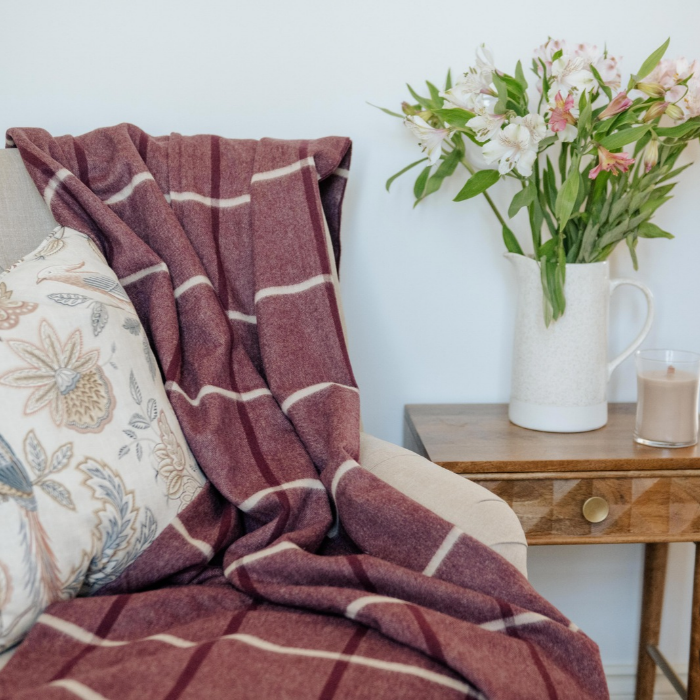 Nithsdale Heather purple pure cashmere blanket in plum and ivory windowpane check with twisted fringe is draped over a beige armchair next to a floral pillow, with a wooden side table holding a white pitcher of flowers and a candle in the background.