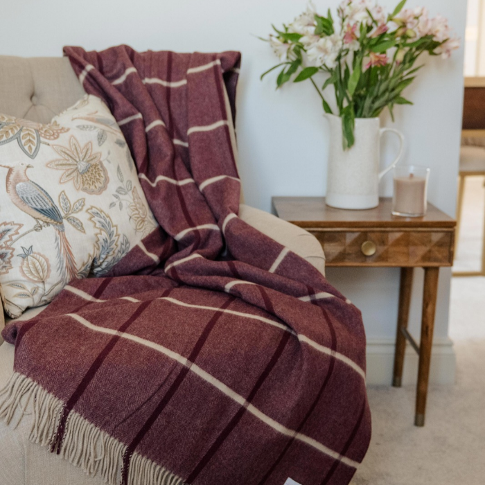 Nithsdale Heather pure cashmere throw in plum and ivory windowpane check with twisted fringe. Draped over an upholstered fabric chair with decorative cushion and side table with vase of flowers and candle