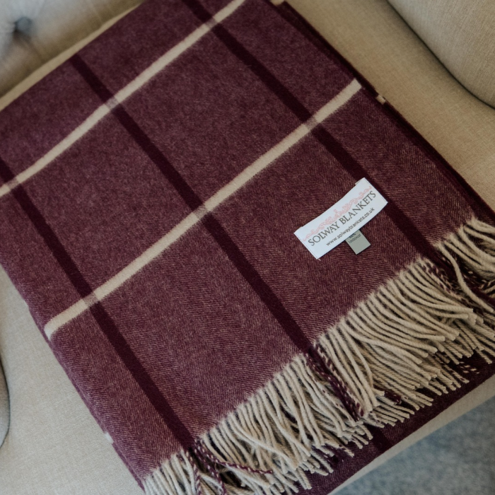 A folded cashmere wool blanket with a deep burgundy base and cream and dark purple plaid stripes, featuring  delicate fringe and a white 'SOLWAY BLANKETS' brand label, displayed on a beige upholstered armchair.