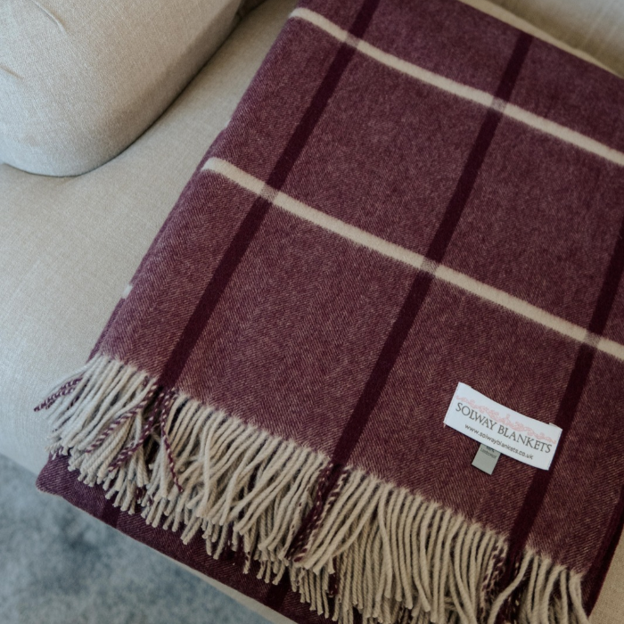 Close-up of fine herringbone weave and damson and ivory windowpane check on plum cashmere draped over a light beige sofa. The blanket features a deep burgundy base with cream and purple check and twisted fringe edge.