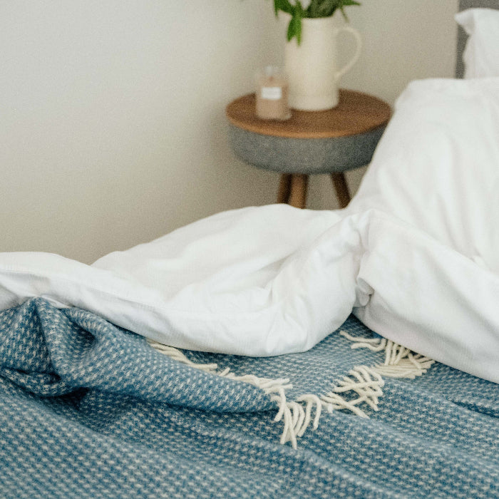 Ocean blue waffle textured wool blanket with white bedding corner folded over featuring a nightstand with plant and candle in the background