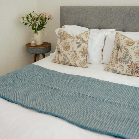 Ocean blue textured wool throw draped over white furnished bed with floral pillows, next to a side table with a vase of flowers.