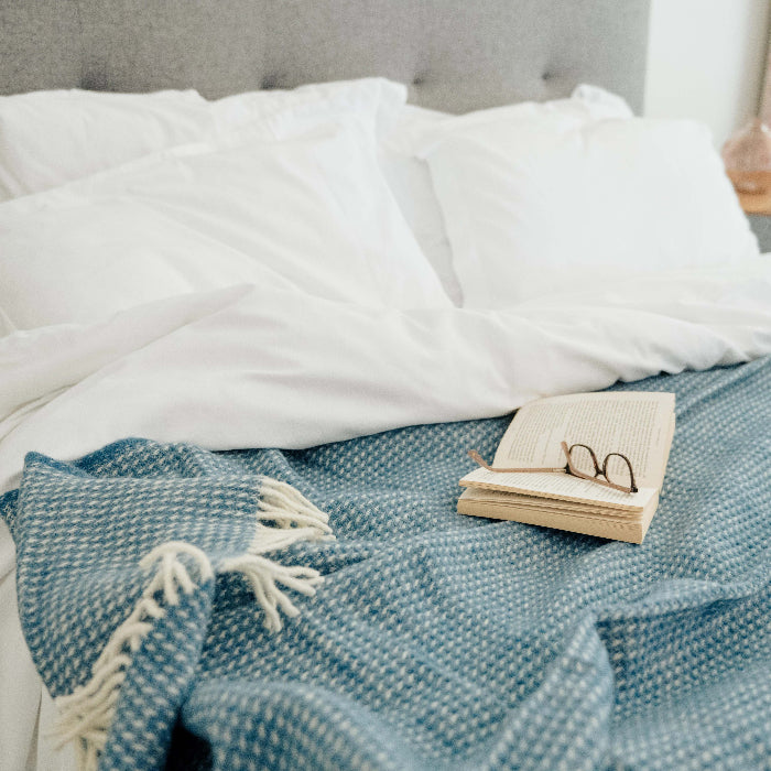 Bed with white pillows with an ocean blue textured wool blanket on top, featuring an open book and a pair of glasses.