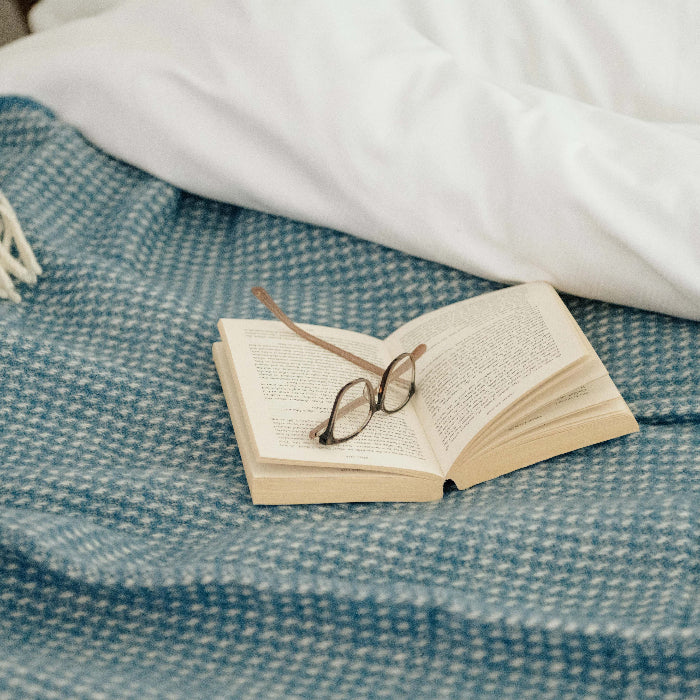 Ocean blue waffle textured wool throw draped over white bedding with an open book and glasses on top