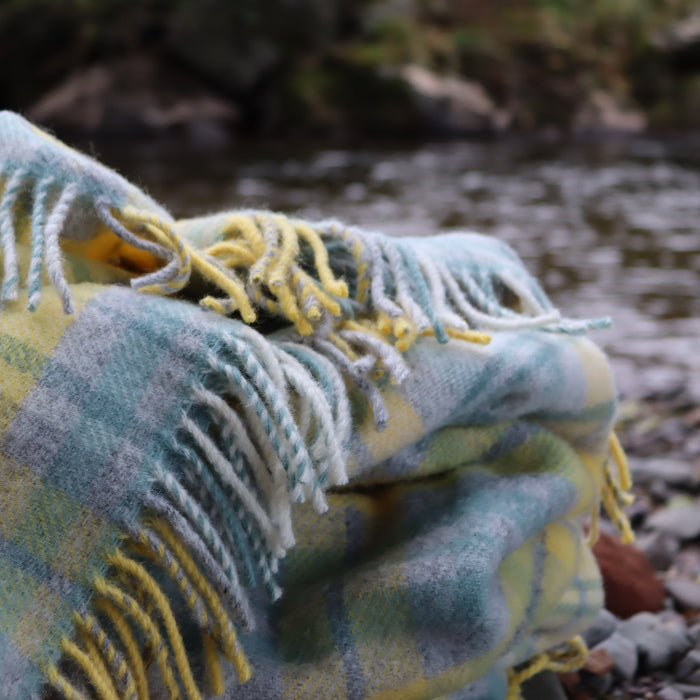Colourful plaid wool blanket with green, grey, white and yellow colourings with fringe on a rocky shore with water in the background