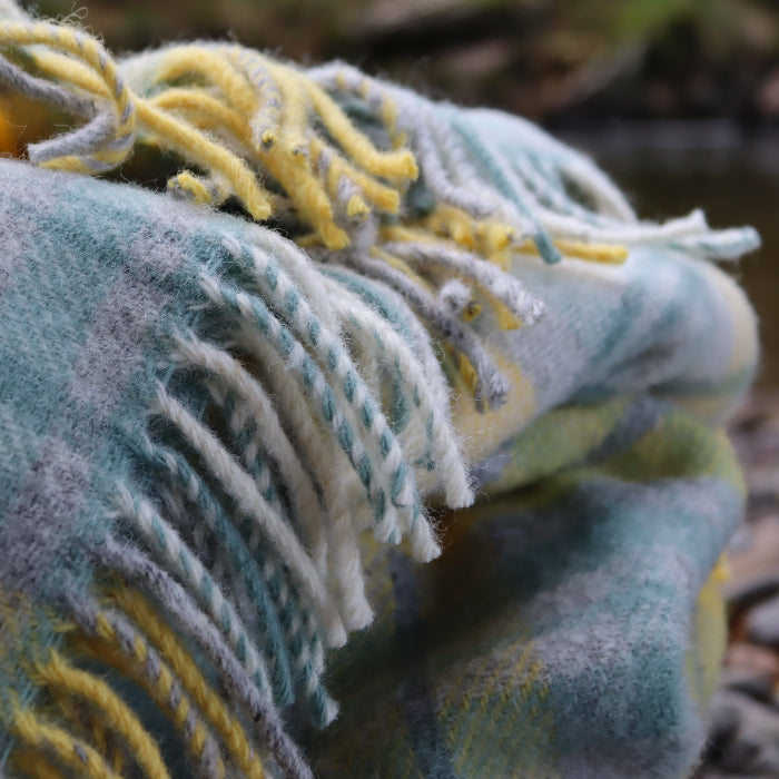 Close up of a sea green, grey, yellow and warm white plaid wool blanket with fringe, set on a stone ground with a river in the background. 