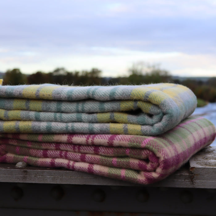 Stack of colourful checkered wool blankets - one with green, grey, white and yellow and the other with pink, purple and green colours - on a wooden bridge with a scenic background