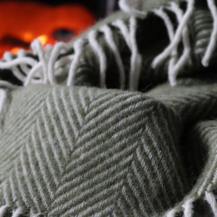 Olive green textured herringbone style wool blanket with fringe against a blurred background of a fire.