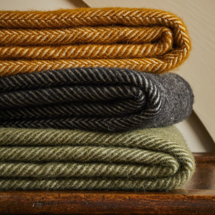 Stack of folded woolen herringbone style blankets in mustard, charcoal, and green on a wooden surface.