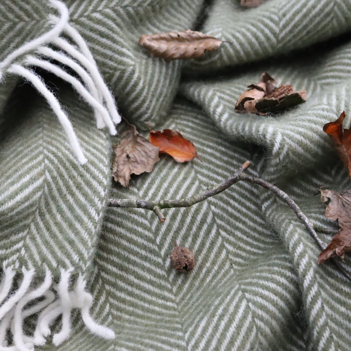 Olive green herringbone wool blanket with white fringe on a surface with leaves