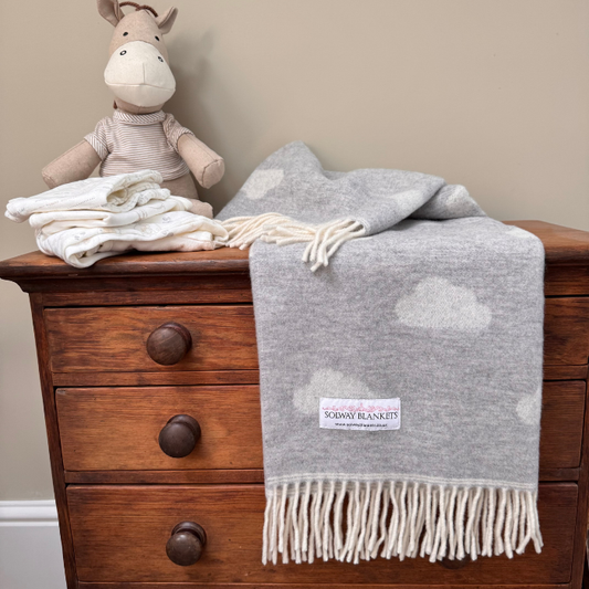A light grey merino wool blanket with off white clouds woven in and fringe, draped over a wooden chest of drawers, with a folded baby clothes and a plush toy on top.