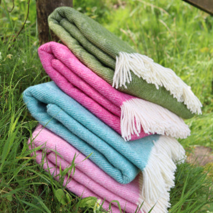 Stack of colourful blankets on a grassy background including the Pale Pink Herringbone Shetland Wool Blanket