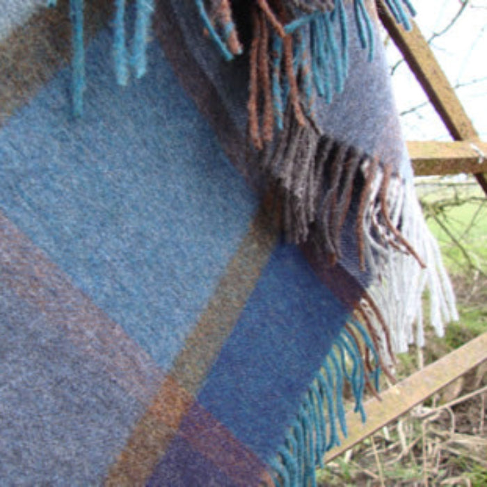 Plaid blanket featuring colors of indigo, teal, denim, rust and grey with fringes draped over a wooden gate outdoors