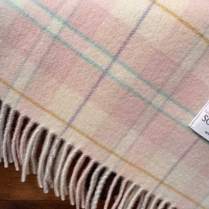 A close-up of a soft woven  cashmere babies receiving blanket with a pastel plaid pattern and fringed edge, laid on a wooden surface. The blanket features a mix of pink, cream, light blue, and yellow colours, British made.