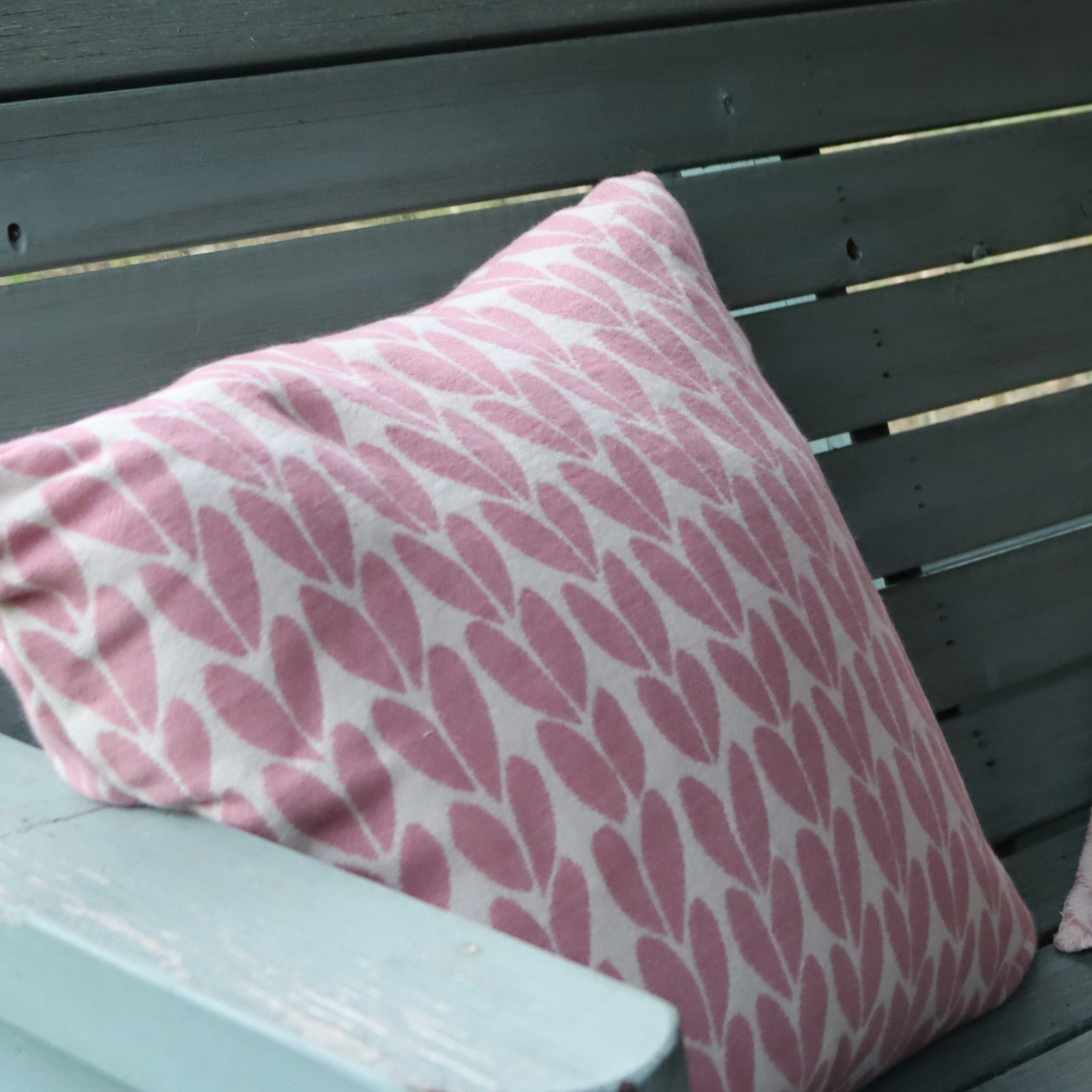 A square throw pillow with a pink and white botanical sycamore leaf pattern, resting on a wooden slatted bench.