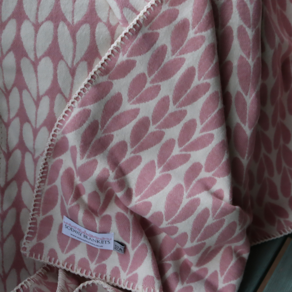 A close-up of a folded throw blanket with a dusty pink and cream leaf pattern, showing its blanket stitched edge and a brand label that reads 'Solway Blankets'.