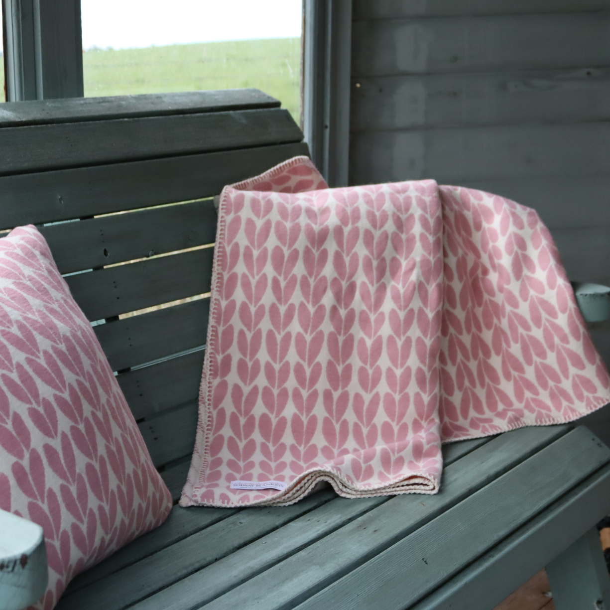 A pink and white natural leaf patterned soft cotton blanket and matching cushion, displayed on a soft green painted wooden bench in summerhouse.
