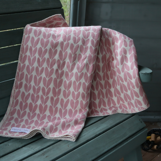A folded pink and off-white leaf patterned blanket or throw with a small visible label, draped over a weathered green wooden bench.