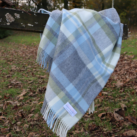 Plaid duck egg blue, grey and green blanket throw with fringes hanging on a wooden bench in an outdoor setting with grass and leaves.