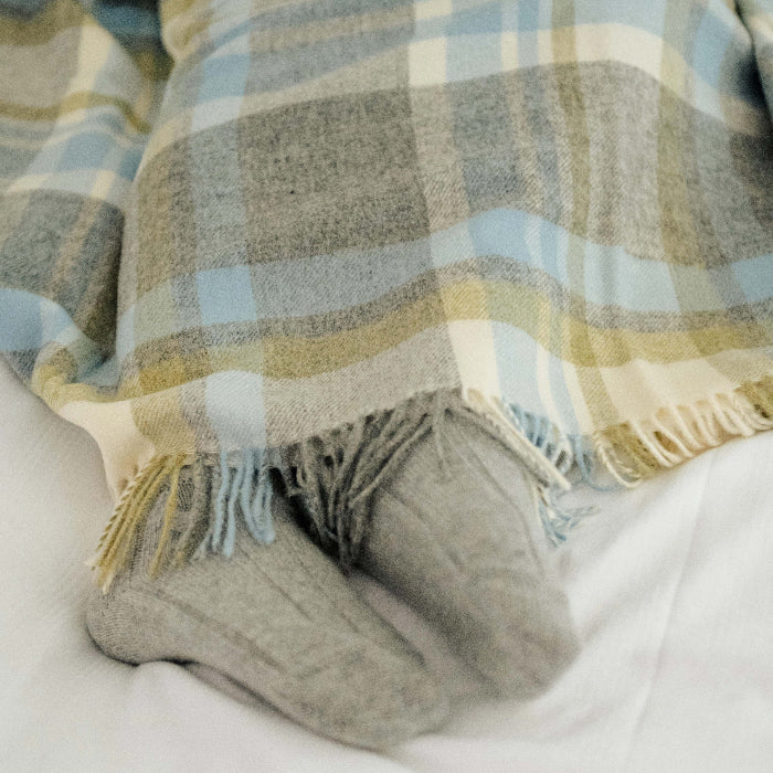 Plaid duck egg blue, grey and green blanket throw draped over a person wearing gray socks on a white bed