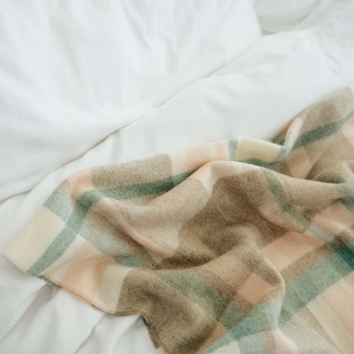 Plaid blanket of cream, beige, soft pink and mute jade colors draped over a white bed